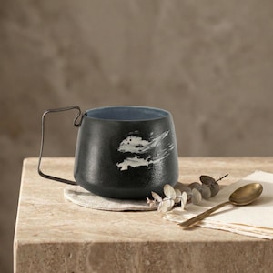 May include: A black ceramic mug with a distinctive metal handle and a blue interior. The mug has a white abstract design on the exterior. It rests on a beige coaster, with a gold spoon and sprig of greenery nearby.