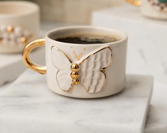 May include: A white ceramic coffee cup with a gold handle and a raised butterfly design. The butterfly is white with gold accents.