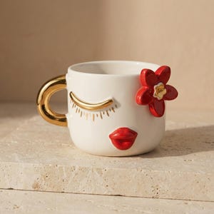 May include: A white ceramic mug with a gold handle and accents. The mug is decorated with a closed eye, red lips, and a red flower. The mug is sitting on a light-colored surface.