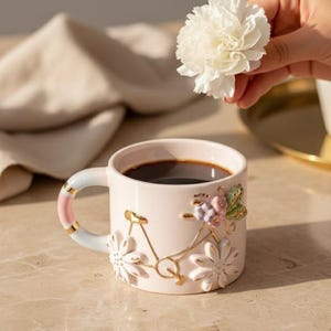 Op de afbeelding: Een lichtroze keramische koffiemok met een fietsontwerp, gevuld met donkere koffie. De mok heeft een witte en roze handgreep met gouden accenten en is versierd met witte bloemen en een gouden fiets. Een witte bloem wordt in de buurt gehouden.