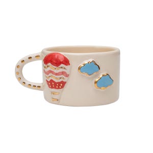 May include: A white ceramic mug with a red, pink, and gold hot air balloon design and two blue clouds with gold outlines.