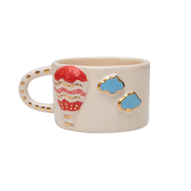 May include: A white ceramic mug with a red, pink, and gold hot air balloon design and two blue clouds with gold outlines.