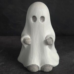 May include: A small, white ghost figurine with a simple design. The ghost has two eye holes and small, rounded arms and feet. The figurine is made of a textured material and is a Halloween decoration.