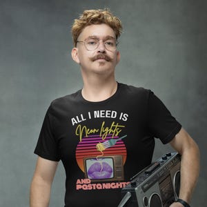 Puede incluir: Una camiseta negra con el texto "All I need is Neon lights and Pasta nights" en blanco. El texto está rodeado por un gráfico retro de un televisor con un labio morado y un tenedor con espaguetis.