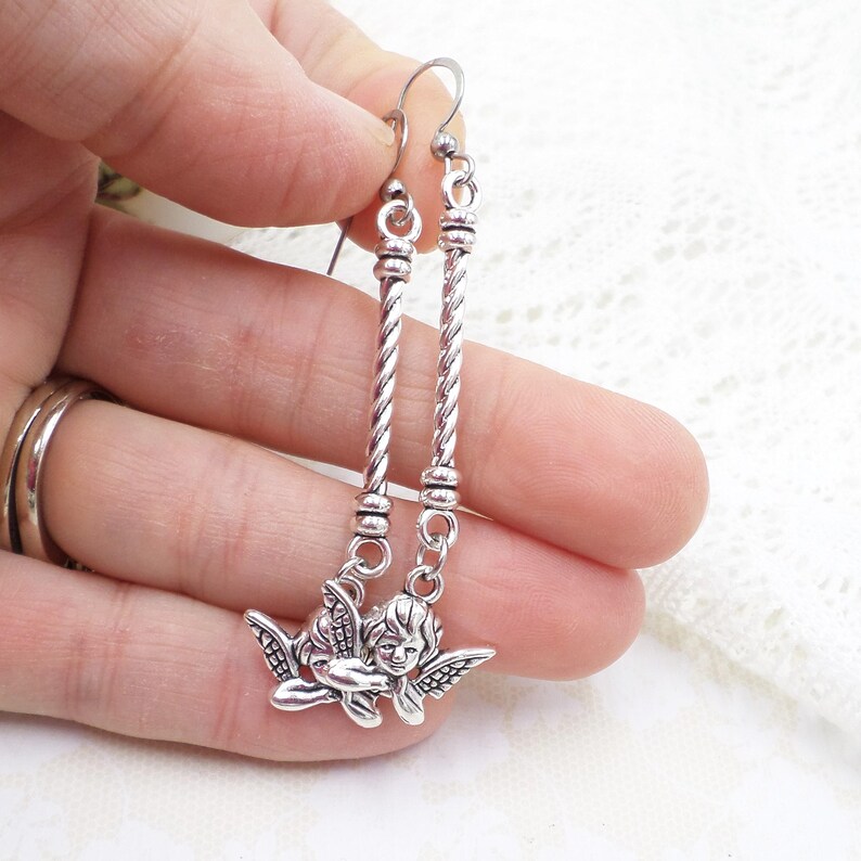 May include: Silver-toned dangle earrings featuring a cherub charm. The earrings have a twisted rope design and a small cherub pendant with detailed wings. The earrings are held up by a silver hook.