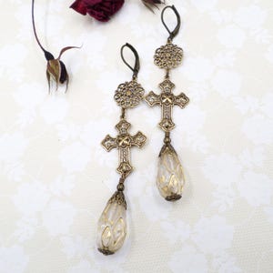 May include: A pair of antique-style earrings with a cross design. The earrings are gold-colored with intricate detailing, featuring crosses and teardrop-shaped pendants with gold accents. The earrings are displayed on a white surface.