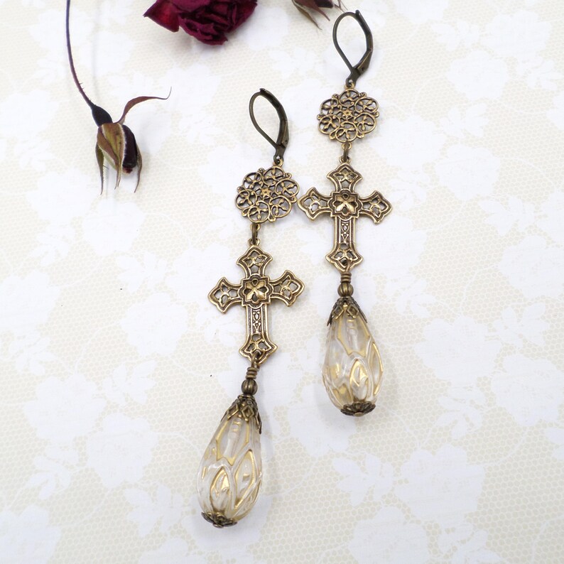 May include: A pair of antique-style earrings with a cross design. The earrings are gold-colored with intricate detailing, featuring crosses and teardrop-shaped pendants with gold accents. The earrings are displayed on a white surface.