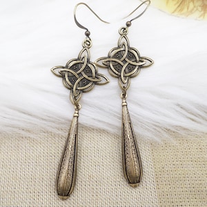 May include: A pair of gold-toned earrings with a Celtic knot design and a teardrop-shaped dangle. The earrings are made of metal and have a polished finish.