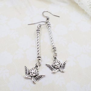 May include: Silver-toned dangle earrings featuring cherub charms. The earrings have a long, twisted bar design with a small cherub pendant at the bottom. The cherubs have detailed wings and facial features. The earrings are displayed on a textured, off-white surface.