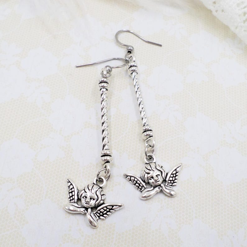 May include: Silver-toned dangle earrings featuring cherub charms. The earrings have a long, twisted bar design with a small cherub pendant at the bottom. The cherubs have detailed wings and facial features. The earrings are displayed on a textured, off-white surface.