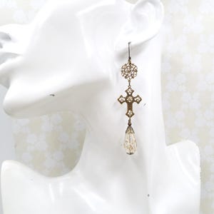 May include: Antique gold-tone earrings with a cross and teardrop design. The earrings feature a filigree cross pendant adorned with small crystals and a pear-shaped, iridescent bead. The top has a circular filigree design and a hook closure.