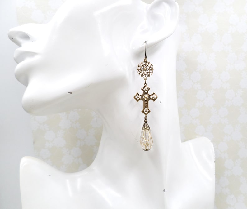 May include: Antique gold-tone earrings with a cross and teardrop design. The earrings feature a filigree cross pendant adorned with small crystals and a pear-shaped, iridescent bead. The top has a circular filigree design and a hook closure.