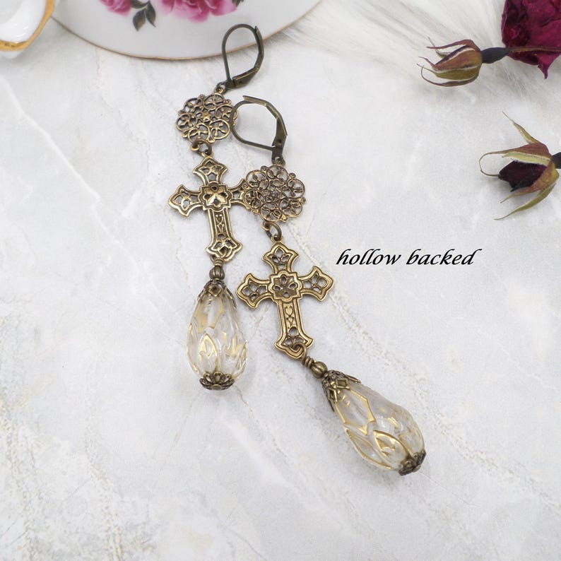 May include: A pair of antique gold-colored earrings. Each earring features a cross pendant, a decorative floral element, and a teardrop-shaped bead. The text "hollow backed" is visible in the image.