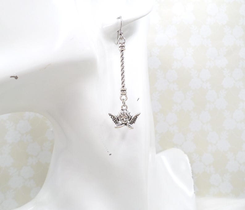May include: A silver-toned earring featuring a cherub charm with detailed wings and a delicate face. The earring hangs from a twisted metal bar, showcasing a classic design. The earring is displayed on a white surface.
