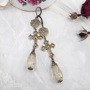 May include: Antique brass-toned earrings featuring ornate crosses and teardrop-shaped clear glass pendants. The earrings have a vintage aesthetic, with intricate filigree details and a hook closure. The clear glass pendants have a delicate, etched design.