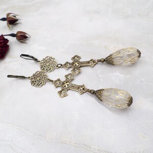 May include: A pair of antique brass-toned earrings. Each earring features a filigree design, a cross, and a teardrop-shaped, translucent bead. The earrings have a lever-back closure and are displayed on a marble surface.