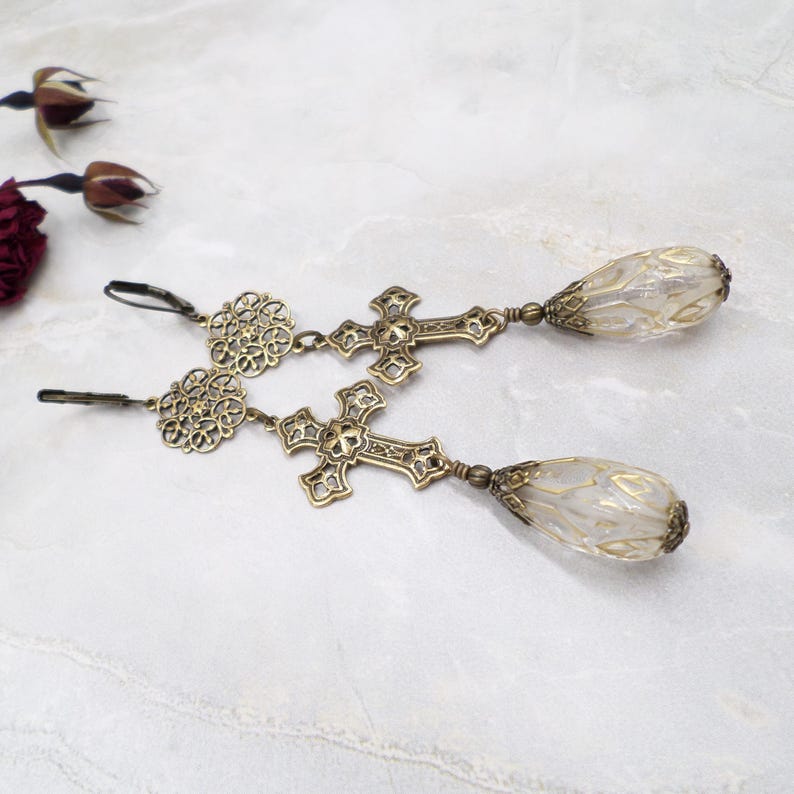 May include: A pair of antique brass-toned earrings. Each earring features a filigree design, a cross, and a teardrop-shaped, translucent bead. The earrings have a lever-back closure and are displayed on a marble surface.