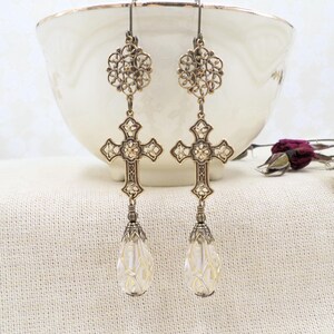 May include: A pair of antique-style earrings with a cross design. The earrings feature a filigree floral design at the top, a detailed cross pendant, and a teardrop-shaped crystal at the bottom. The earrings are gold-toned and hang from a hook.