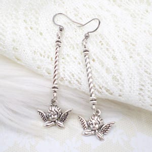 May include: Silver-toned dangle earrings featuring a cherub design. Each earring has a long, twisted bar with decorative beads and a small cherub pendant with detailed wings. The earrings are suspended from a simple hook.