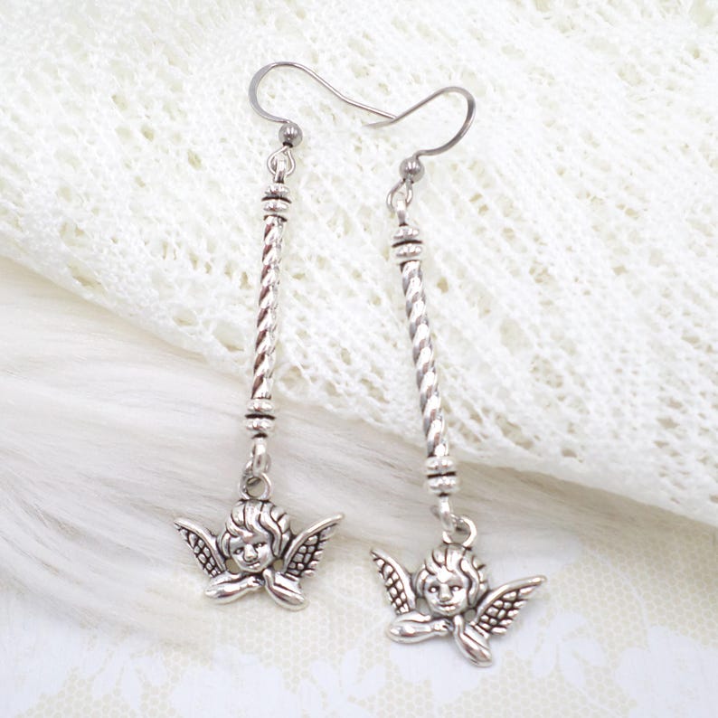 May include: Silver-toned dangle earrings featuring a cherub design. Each earring has a long, twisted bar with decorative beads and a small cherub pendant with detailed wings. The earrings are suspended from a simple hook.