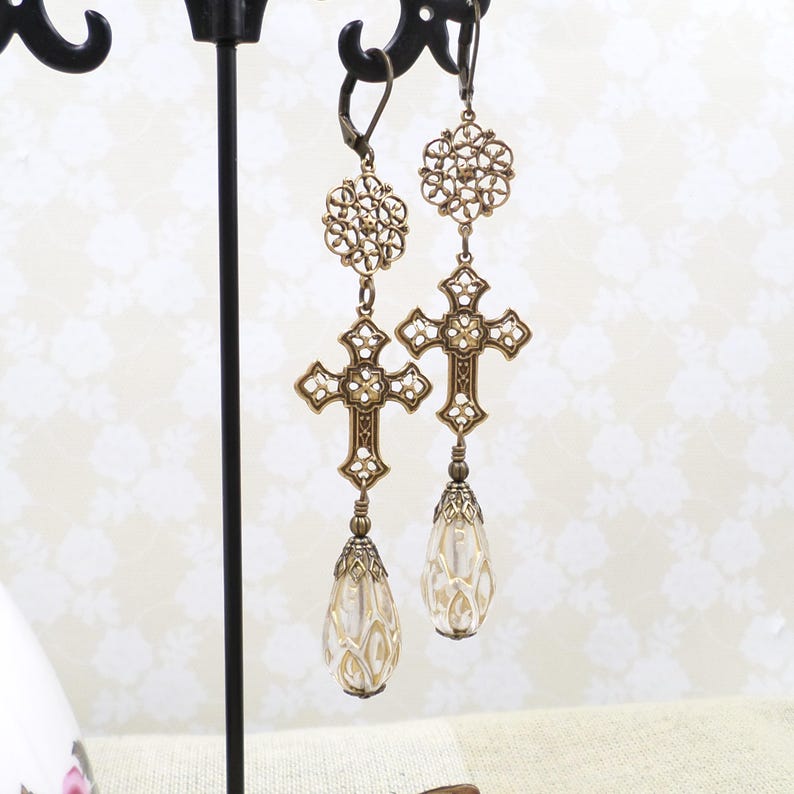 May include: A pair of antique gold-colored earrings. Each earring features a filigree cross design with a teardrop-shaped, clear, beaded charm. The earrings are suspended from a decorative floral element and are displayed on a black stand.