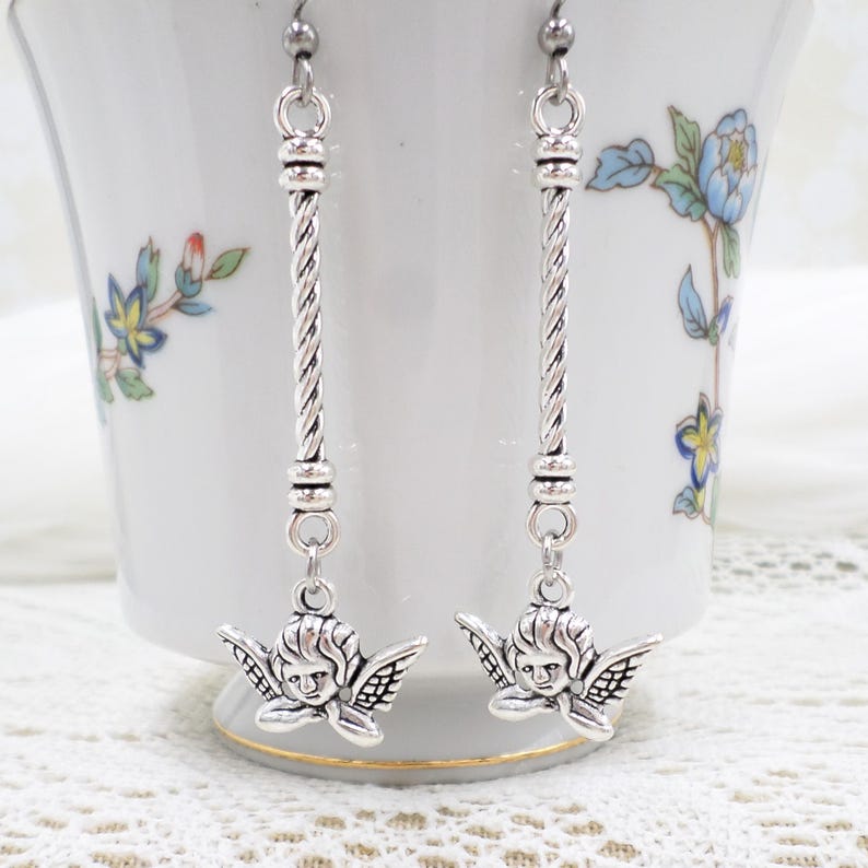 May include: Silver-toned dangle earrings featuring a cherub design. The earrings have a long, twisted bar with a cherub pendant at the bottom. The cherubs have detailed wings and facial features. The earrings are displayed against a white background.
