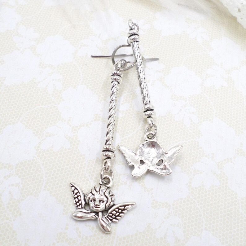 May include: Silver-toned toggle clasp with two dangling charms. One charm is an angel with detailed wings and face, the other is a stylized mask. The clasp has a twisted rope design.