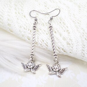 May include: Silver-toned dangle earrings featuring a cherub design. Each earring has a long, twisted bar with a cherub pendant at the bottom. The cherubs have detailed wings and smiling faces. The earrings are displayed on a white, textured background.