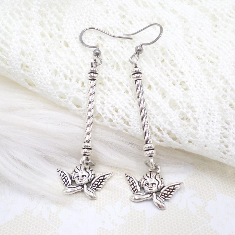 May include: Silver-toned dangle earrings featuring a cherub design. Each earring has a long, twisted bar with a cherub pendant at the bottom. The cherubs have detailed wings and smiling faces. The earrings are displayed on a white, textured background.