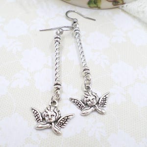 May include: Silver-toned dangle earrings featuring cherub charms. Each earring has a long, twisted bar with a cherub pendant at the bottom. The cherubs have detailed wings and facial features. The earrings are designed to be worn with pierced ears.