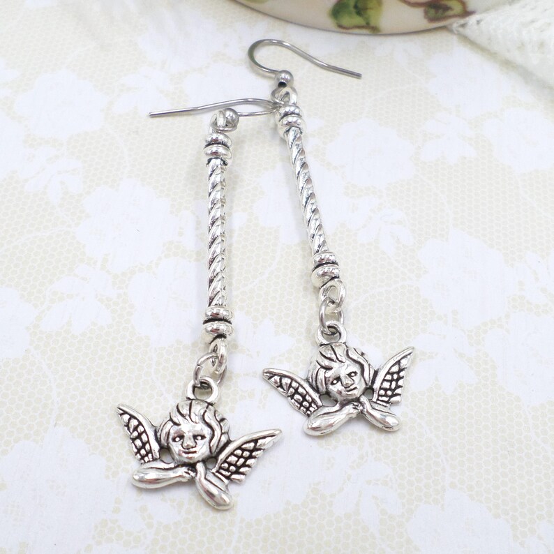May include: Silver-toned dangle earrings featuring cherub charms. Each earring has a long, twisted bar with a cherub pendant at the bottom. The cherubs have detailed wings and facial features. The earrings are designed to be worn with pierced ears.