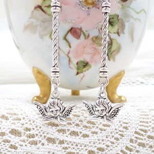 May include: Silver-toned dangle earrings featuring cherub charms. The earrings have a twisted rope design and small decorative beads. The cherub charms have detailed wings and facial features. The earrings are displayed on a white lace surface.
