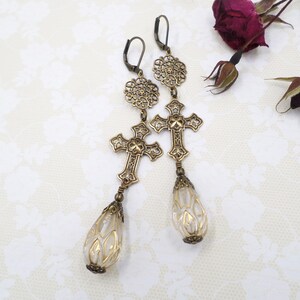 May include: A pair of antique brass-toned earrings. Each earring features a filigree flower, a cross pendant, and a teardrop-shaped bead with a gold floral design. The earrings have a vintage, gothic aesthetic.