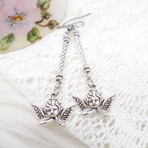 May include: Silver-toned dangle earrings featuring cherub charms. The earrings have a twisted rope design and small angel pendants with detailed wings and serene expressions. The earrings are displayed on a white lace surface.