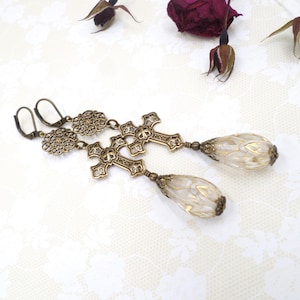 May include: A pair of antique-style earrings featuring bronze-colored crosses and floral accents. The earrings have teardrop-shaped, off-white and gold-colored glass beads. The earrings are designed with a lever-back closure.