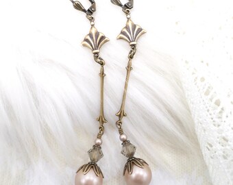 Pode incluir: Um par de brincos pendurados em estilo antigo. Os brincos apresentam um metal de cor bronze com um topo em forma de leque, uma haste longa e fina e uma grande pérola redonda cor de rosa na parte inferior. Pequenos cristais transparentes e pérolas menores também estão incluídos.