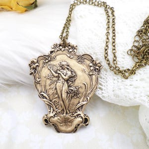 May include: Antique gold-tone pendant necklace featuring a detailed Art Nouveau design. The pendant showcases a woman playing a harp, surrounded by floral motifs. The necklace has a delicate chain and a decorative clasp.