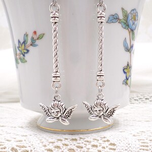 May include: Silver-toned dangle earrings featuring cherub charms. The earrings have a twisted bar design and are suspended from a simple hook. The cherub charms have detailed wings and facial features. The earrings are displayed against a white background.