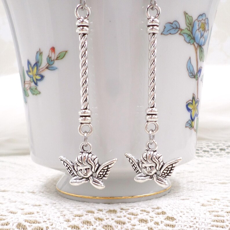 May include: Silver-toned dangle earrings featuring cherub charms. The earrings have a twisted bar design and are suspended from a simple hook. The cherub charms have detailed wings and facial features. The earrings are displayed against a white background.