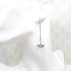 May include: Silver-toned dangle earring featuring a cherub charm. The earring has a twisted rope design and a small cherub pendant with detailed wings. The earring is displayed on a white mannequin head.