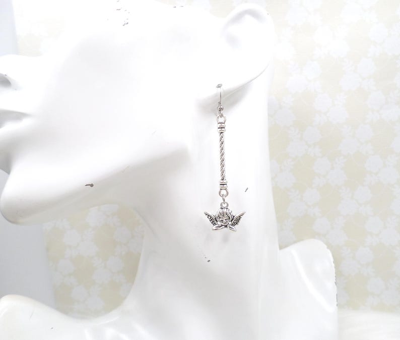 May include: Silver-toned dangle earring featuring a cherub charm. The earring has a twisted rope design and a small cherub pendant with detailed wings. The earring is displayed on a white mannequin head.