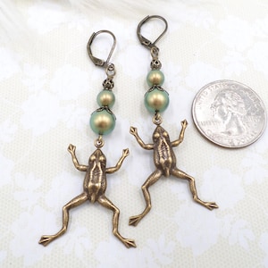 May include: A pair of bronze-toned frog earrings. Each earring features a frog charm with outstretched arms, suspended from two green and gold beads and a decorative top. The earrings have lever-back closures.