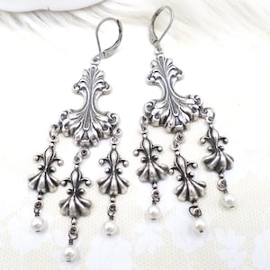 May include: A pair of silver chandelier earrings. Each earring features an ornate, stylized floral design with dangling elements. Small, white faux pearls are suspended from the bottom of each earring, adding a touch of elegance to the design.