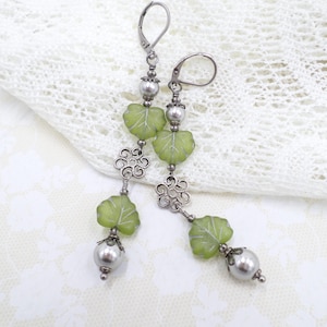 May include: Long dangle earrings featuring green leaf-shaped beads with white veining, silver-tone filigree accents, and gray pearl-like beads. The earrings have lever-back closures and are displayed on a white lace background.