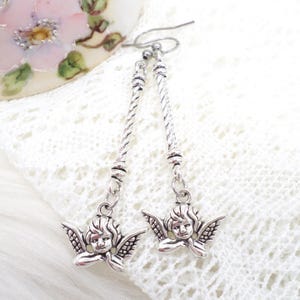 May include: Silver-toned dangle earrings featuring cherub charms. The earrings have a twisted rope design on the drop and a small loop at the top for the ear wire. The cherub charms have detailed wings and a serene expression.