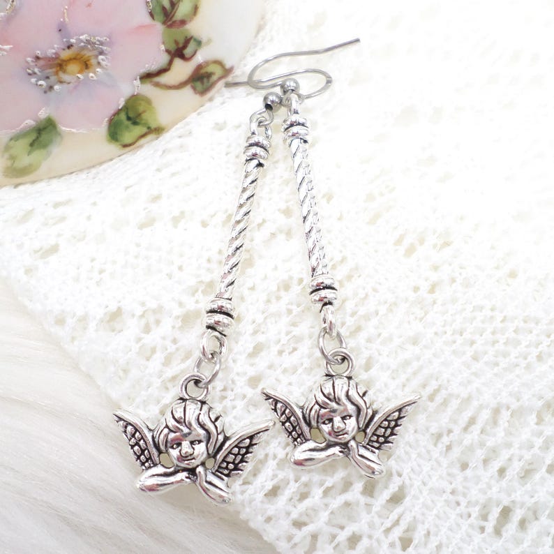 May include: Silver-toned dangle earrings featuring cherub charms. The earrings have a twisted rope design on the drop and a small loop at the top for the ear wire. The cherub charms have detailed wings and a serene expression.