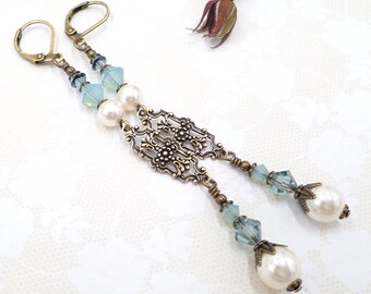 May include: A pair of long dangle earrings featuring antique bronze lever-back hooks, faceted light blue crystal beads, and round white faux pearls. The central design consists of an intricate, openwork bronze filigree component.