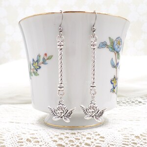 May include: Silver-toned dangle earrings featuring a cherub design. The earrings have a long, twisted bar with a cherub pendant at the bottom. The cherub has detailed wings and a smiling face. The earrings are displayed in front of a white teacup with floral designs.