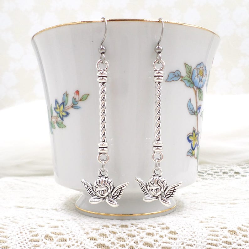 May include: Silver-toned dangle earrings featuring a cherub design. The earrings have a long, twisted bar with a cherub pendant at the bottom. The cherub has detailed wings and a smiling face. The earrings are displayed in front of a white teacup with floral designs.