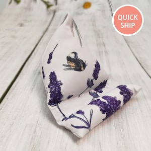 May include: A white lavender and bumblebee print fabric pillow with a triangular shape. The pillow is folded in half and has a flat surface. The pillow is on a white wooden surface.
